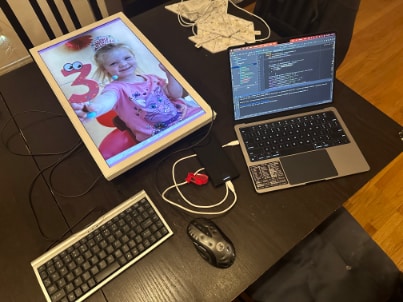 A lit EO1 lying flat on a table showing a child's birthday photo, beside an open laptop, a keyboard and a mouse.