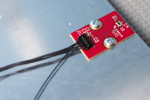 Macro of the small red ambient-light sensor board screwed to brushed metal, with four thin wires leaving frame.